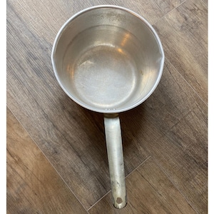 May include: A vintage aluminum saucepan with a long handle. The pan has a rounded shape with a pouring spout and a silver finish. The handle is riveted to the pan and has a hole at the end for hanging.