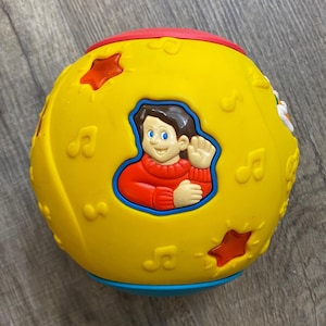 May include: A yellow spherical toy featuring a raised relief of a person in a red sweater waving. The surface is decorated with embossed musical notes and star shapes, with a red top section and a blue base section, creating a colorful design.