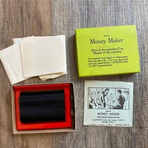 May include: A vintage Adams Money Maker toy set featuring a bright green box with black text, a red plastic roller device with black rollers, several blank white paper strips, and a printed instruction sheet with black and white illustrations.
