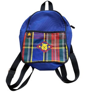 May include: A blue backpack with black straps and a top handle. The front pocket features a plaid pattern in red, green, yellow, and blue, with a Winnie the Pooh patch. A yellow star is on the upper left corner.