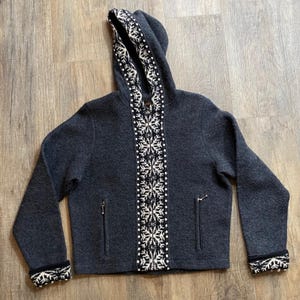 May include: A charcoal gray hooded jacket featuring a central vertical panel and cuffs decorated with a white snowflake pattern. The garment includes a full-length front zipper, a hood, and two zippered pockets on the lower front section.