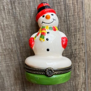 May include: A ceramic snowman figurine trinket box with a glossy white finish. The snowman wears a red hat, red mittens, and a multi-colored striped scarf. It features a black-dotted face and buttons, resting on a green base with a metal hinge.