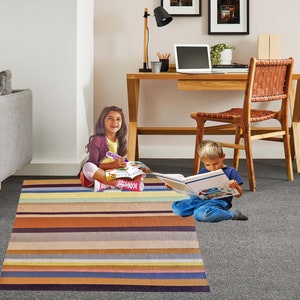 May include: Two children are sitting on a striped rug, one reading a book and the other drawing in a notebook. The rug has a variety of colors, including brown, beige, pink, and blue.