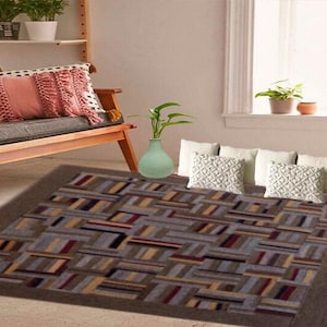 May include: A rectangular area rug with a geometric pattern in shades of brown, gray, beige, and burgundy. The rug is placed on a light-colored floor. A light green vase with a plant is on the rug. Decorative pillows and a wooden bench are in the background.