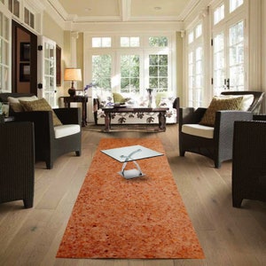 May include: A long, rectangular, orange and brown area rug with a glass coffee table in the center of the rug. The coffee table has a clear glass top and a silver metal base. The rug is in a living room with two wicker chairs and a sofa.