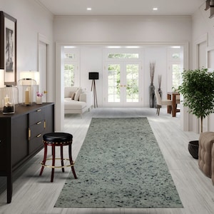 May include: A light gray area rug with a speckled pattern is placed in the middle of a hallway with white walls and wood floors. A black leather stool with gold accents is placed to the left of the rug. A dark brown dresser with gold handles is to the left of the stool. Two lamps with white shades are on top of the dresser. A small green plant is in a pot on the floor next to the dresser. A small green plant is in a pot on the floor to the right of the rug. A brown ottoman with a tufted design is to the right of the rug.