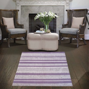 May include: A striped area rug with purple and white stripes in a living room setting with two wicker chairs and a white ottoman in front of a fireplace.