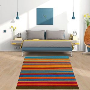 May include: A colorful striped rug with orange, blue, red, and yellow stripes in a bedroom setting. The rug is in front of a bed with gray bedding and blue accents.