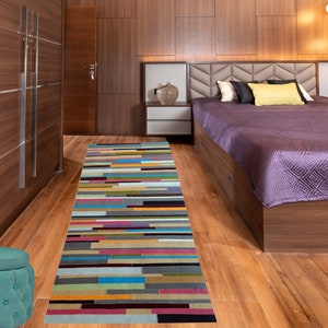 May include: A colorful striped rug with a teal, pink, and yellow color scheme. The rug is laid out in a hallway with a wooden floor and a bedroom with a purple bedspread in the background.