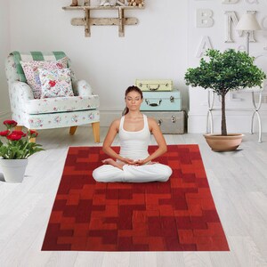 May include: A woman sits in a lotus position on a red and brown patterned yoga mat. The mat has a geometric design. The woman is wearing a white tank top and white pants. She is meditating with her eyes closed.
