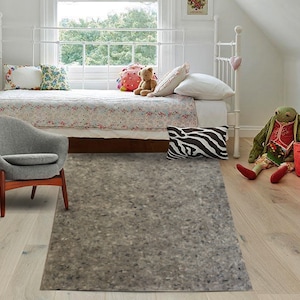 May include: A gray rug with a speckled texture, placed in a bedroom with a white bed, a gray armchair, and a wooden floor.