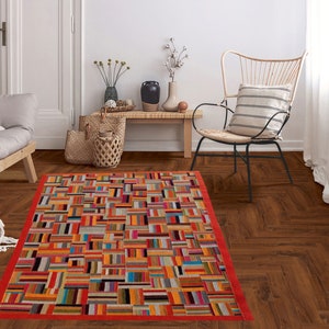 May include: A colorful patchwork rug with a red border, placed on a hardwood floor in a living room setting. The rug features a geometric pattern of squares and rectangles in various shades of orange, yellow, blue, green, and brown.