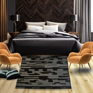 May include: A black and gray geometric patterned rug with a modern design. The rug is placed in a bedroom setting with a bed, two chairs, and a wooden floor.