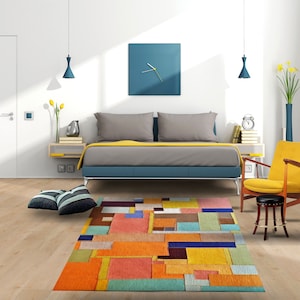 May include: A colorful geometric patterned rug with shades of orange, yellow, blue, green, and brown. The rug is in a living room setting with a bed, a chair, and a side table.