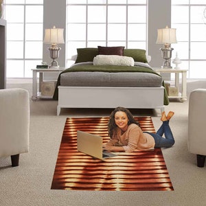 May include: A woman is sitting on a brown and orange striped rug with a laptop computer. She is wearing a light pink sweater and blue jeans. The rug is in a living room with a white couch and a white bed in the background.