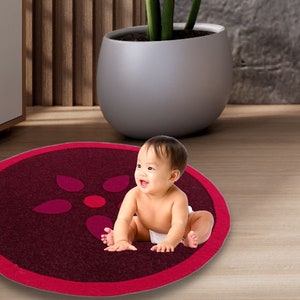 May include: A baby sits on a round, burgundy play mat with a pink border and a pink flower design.