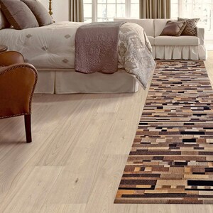May include: A long, rectangular brown and black area rug with a geometric pattern. The rug is laid out on a light wood floor in a bedroom setting.