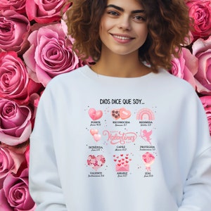 May include: White sweatshirt with a pink and white graphic design that says "Dios dice que soy..." and features a heart with the words "Valentines Day" in pink. The graphic also includes several pink hearts with bible verses and words like "fuerte", "intrepida", "capaz", "redimida", "protegida", "valiente", "amable", and "leal".