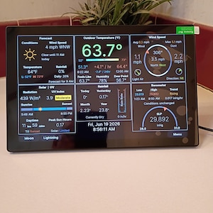May include: A rectangular digital weather station display screen showing real-time data including outdoor temperature of 63.7 degrees Fahrenheit, wind speed, barometric pressure, solar radiation, UV index, and rainfall statistics with a date of June 19, 2026.