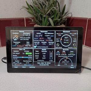 Puede incluir: Estación meteorológica digital negra que muestra las condiciones climáticas actuales, incluyendo temperatura, velocidad del viento y pronóstico. La pantalla muestra lecturas en Fahrenheit y mph. Una planta en el fondo.