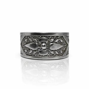 May include: A silver-colored ring with an intricate floral design. The ring features a central flower motif surrounded by smaller leaf-like details and a textured background. The ring is displayed on a reflective surface.