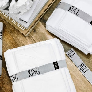 May include: White bed sheets are neatly stacked and secured with gray bands labeled "FULL" and "KING." The bands feature black text and a small logo. The sheets are on a wooden surface, with books and flowers in the background.