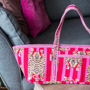 Puede incluir: Un bolso tote vibrante con un estampado de tigre repetido sobre un fondo a rayas. El bolso presenta una combinaci&oacute;n de rayas rosas y rojas, con un asa a juego. El bolso est&aacute; hecho de un material acolchado.