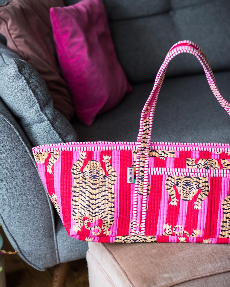 Puede incluir: Un bolso tote vibrante con un estampado de tigre repetido sobre un fondo a rayas. El bolso presenta una combinaci&oacute;n de rayas rosas y rojas, con un asa a juego. El bolso est&aacute; hecho de un material acolchado.
