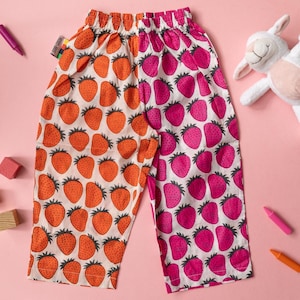 Op de afbeelding: Een broek met elastische tailleband en een tweekleurig ontwerp. De linkerkant is wit met oranje aardbeienprints en de rechterkant is wit met roze aardbeienprints. Aan de taille is een klein stoffen label bevestigd.