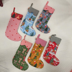 Könnte beinhalten: Sechs Weihnachtssocken mit verschiedenen festlichen Mustern. Designs umfassen rot-weiße Streifen, Skifahrer, Geschenkboxen, Weihnachtsbäume und Schleifen. Jede Socke hat eine gestreifte Manschette und eine Schlaufe zum Aufhängen. Die Socken sind auf einer weißen Oberfläche angeordnet.
