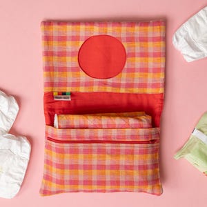 May include: A rectangular fabric pouch featuring an orange and pink gingham pattern with a circular opening on the top flap. The interior is lined with solid red fabric and includes a zippered pocket. A small label reads Merry Brite.