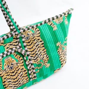 Puede incluir: Un bolso tote acolchado verde con estampado de tigre. El bolso presenta rayas verdes verticales y un patr&oacute;n repetido de tigres en tonos beige y negro. Las asas y el ribete est&aacute;n a rayas en verde, blanco y rosa.