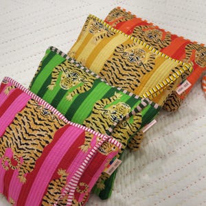 May include: Four colourful zippered pouches with a tiger print design. The pouches are lined up in a row, with the pink pouch at the front and the orange pouch at the back. Each pouch has a white stripe along the edge and a small tag that says "Vicky + Brett".