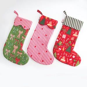 May include: Three festive Christmas stockings in red, green, and white. The green stocking features bows and candy canes. The red stocking has a striped pattern with hearts. The third stocking is red with Christmas trees and gifts.