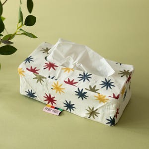 May include: A rectangular fabric tissue box cover in white, featuring a repeating pattern of star-shaped motifs in red, yellow, dark blue, and olive green. A small rectangular tag on the side displays the text MERRY + BRITE in black lettering.