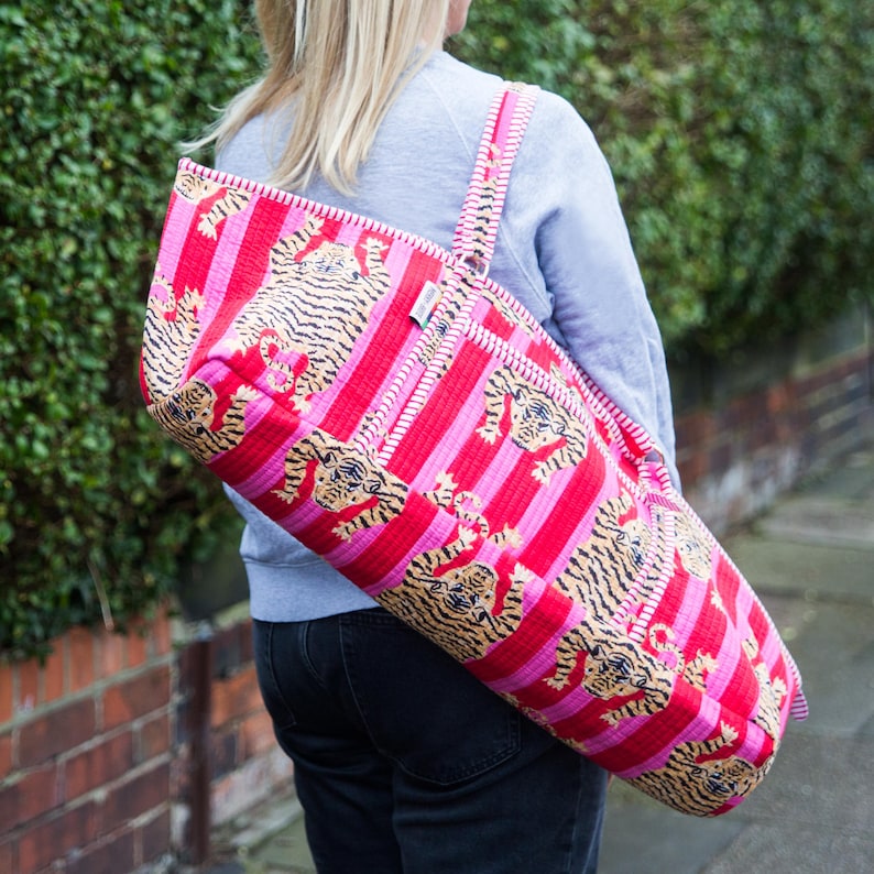 Puede incluir: Una bolsa para esterilla de yoga a rayas rojas y rosas con un estampado de tigre repetido. La bolsa tiene una correa para el hombro y se lleva sobre el hombro. La bolsa est&aacute; hecha de una tela acolchada.