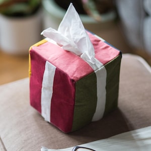 May include: A colourful striped tissue box cover with a white tissue inside. The cover is made of fabric and has a square shape. The box is sitting on a brown surface.