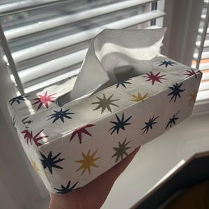 Może przedstawiać: Prostokątne pudełko na chusteczki z białym tłem i powtarzającym się wzorem kolorowych gwiazd w kolorach czerwonym, żółtym, zielonym i niebieskim. Biała chusteczka wystaje z górnego otworu. Pudełko jest trzymane w dłoni.