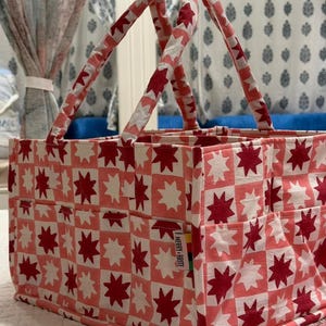 Puede incluir: Un bolso tote rectangular con un patrón repetido de estrellas rojas y blancas sobre un fondo rosa. El bolso tiene dos asas y varios bolsillos exteriores. La etiqueta lateral dice "MERRY + BRITE".