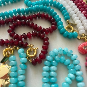 Puede incluir: Una colección de collares de cuentas en varios colores, incluyendo turquesa, blanco y rojo oscuro. Los collares tienen cierres dorados y una variedad de formas de cuentas. La imagen muestra las texturas y variaciones de color de los collares.