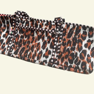 Puede incluir: Un bolso tote rectangular con un estampado de leopardo en negro, marr&oacute;n y blanco. El bolso tiene dos asas y un ribete a rayas blancas y negras. El ribete interior es rojo.