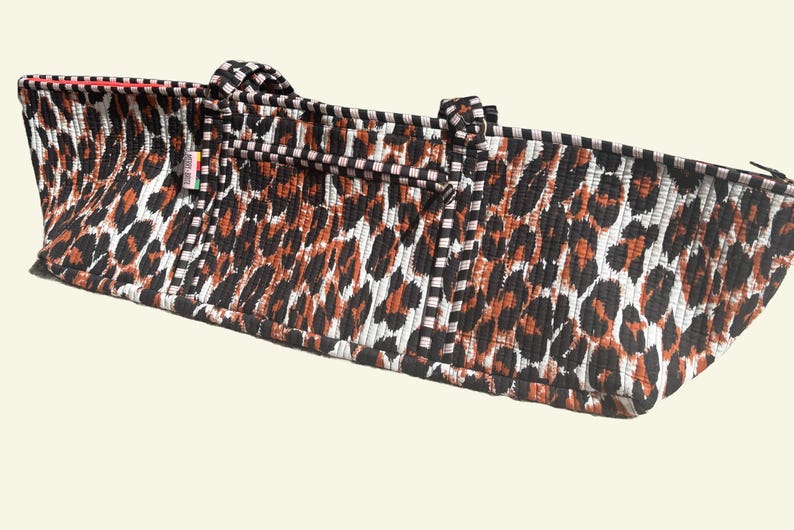 Puede incluir: Un bolso tote rectangular con un estampado de leopardo en negro, marr&oacute;n y blanco. El bolso tiene dos asas y un ribete a rayas blancas y negras. El ribete interior es rojo.
