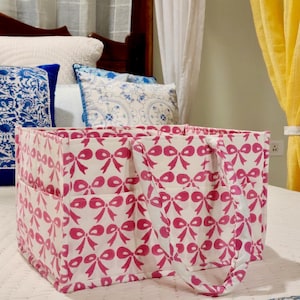 May include: A white canvas tote bag with pink bow print and two handles. The bag has multiple pockets and is ideal for organising and carrying items.