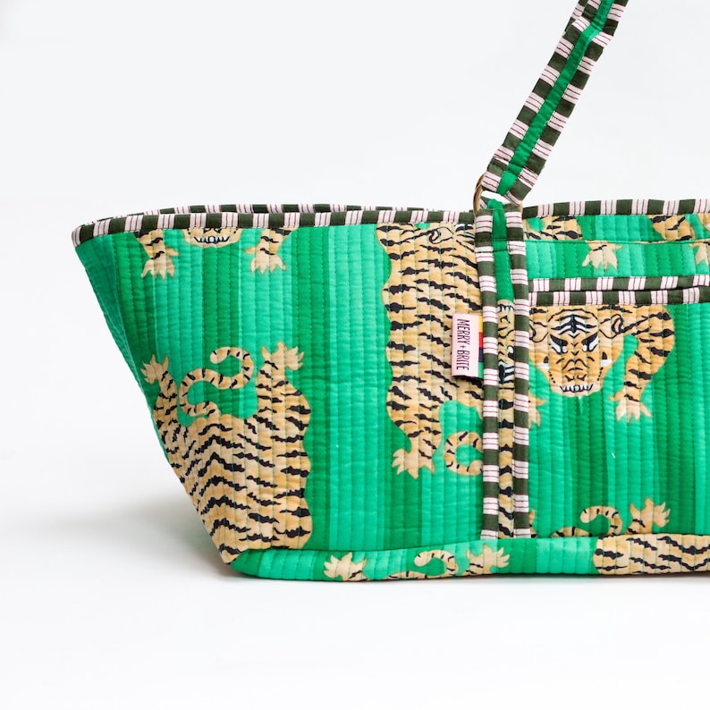 Puede incluir: Bolso tote acolchado verde con estampado de tigre. El bolso presenta rayas verticales y un patr&oacute;n repetido de tigres en tonos beige y negro. Las asas y el ribete tienen un dise&ntilde;o a rayas en verde, blanco y rosa. La etiqueta dice "Merry + Bright".