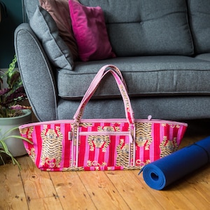 Puede incluir: Una bolsa de yoga rosa y roja con un dise&ntilde;o de estampado de tigre. La bolsa tiene forma rectangular con dos asas y un cierre de cremallera. Una esterilla de yoga azul enrollada descansa en el suelo de madera.
