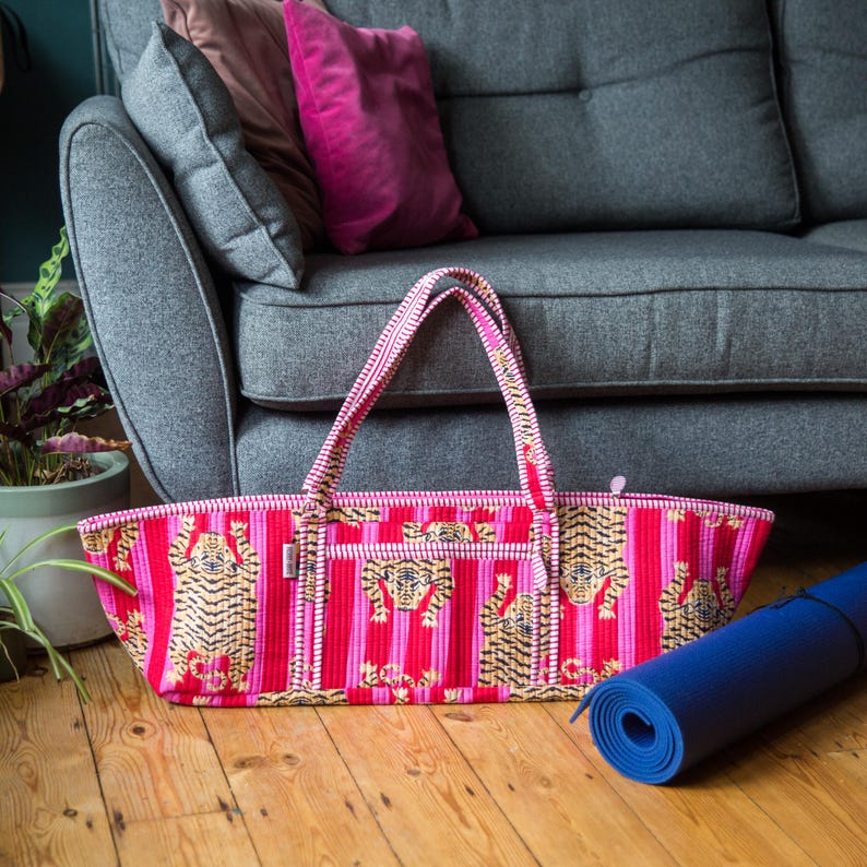 Puede incluir: Una bolsa de yoga rosa y roja con un dise&ntilde;o de estampado de tigre. La bolsa tiene forma rectangular con dos asas y un cierre de cremallera. Una esterilla de yoga azul enrollada descansa en el suelo de madera.