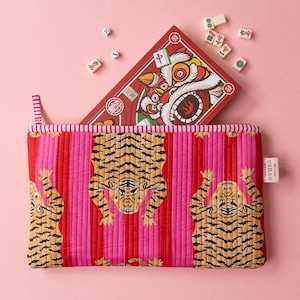 May include: A rectangular quilted pouch featuring a repeating tiger pattern on a background of vertical red and pink stripes. A red box with a lion illustration and several small mahjong tiles are positioned behind the pouch.