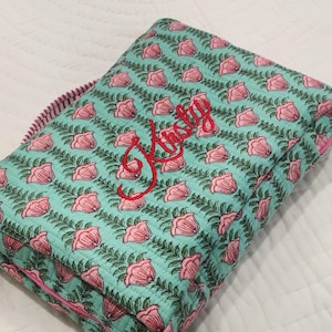 May include: A folded, quilted blanket featuring a floral design of pink roses and green leaves on a teal backdrop. The blanket has a red embroidered name, "Kristy". The edges are trimmed with pink fabric.