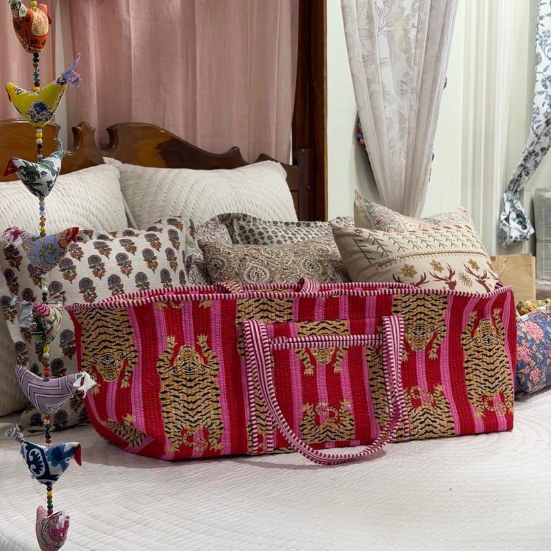 May include: A rectangular quilted tote bag featuring a vibrant pattern of tigers on alternating red and pink vertical stripes. The bag has two matching striped fabric handles and a quilted texture throughout the exterior surface.