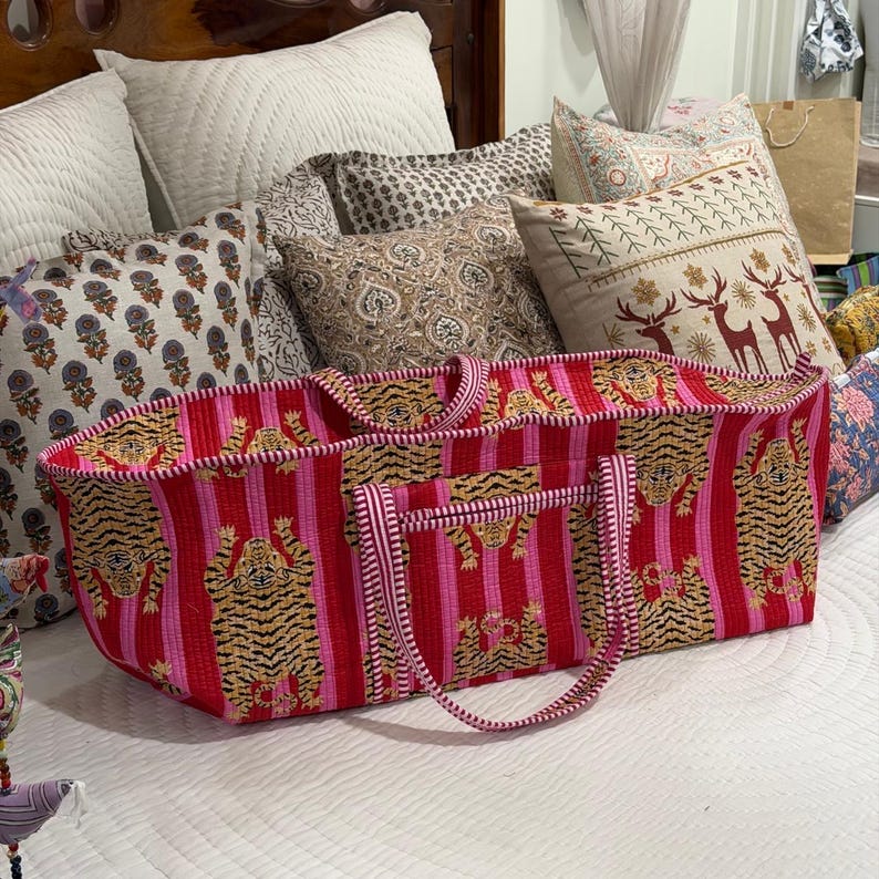 May include: A rectangular quilted tote bag featuring a vibrant pattern of tigers on alternating vertical stripes of bright pink and red. The bag includes two matching striped fabric handles and a soft, stitched texture throughout the exterior.
